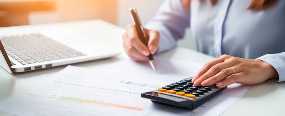 O que é RC contador? Na imagem, mulher fazendo cálculos com calculadora, caneta e papel em alusão atividade do contador, que pode receber proteção financeira por meio de seguro de responsabilidade civil contador