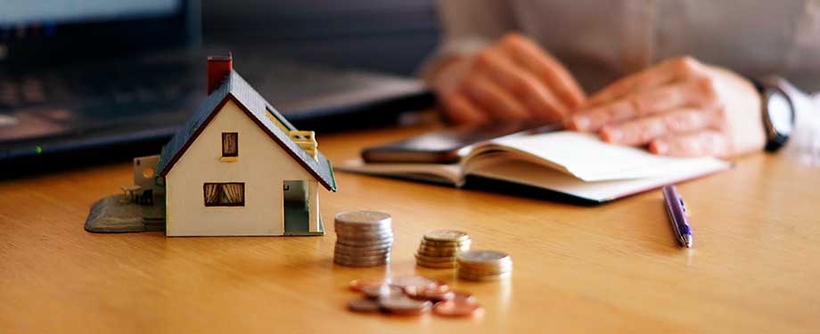 O que cobre o seguro garantia imobiliário? Na imagem, pessoa com caderno, casa em miniatura e moedas representando as possibilidades de cobertura, que podem variar de acordo com o tipo de seguro